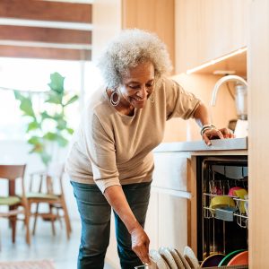 Smiling senior woman keeping plates in dishwasher. Retired elderly female is doing routine chores in kitchen. She is in casuals at home.