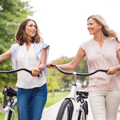 Two mature women in conversation while walking with bicycle at park. Happy beautiful senior women walking in the park with bicycles in a spring time. Friends holding bikes and talking to each other."r